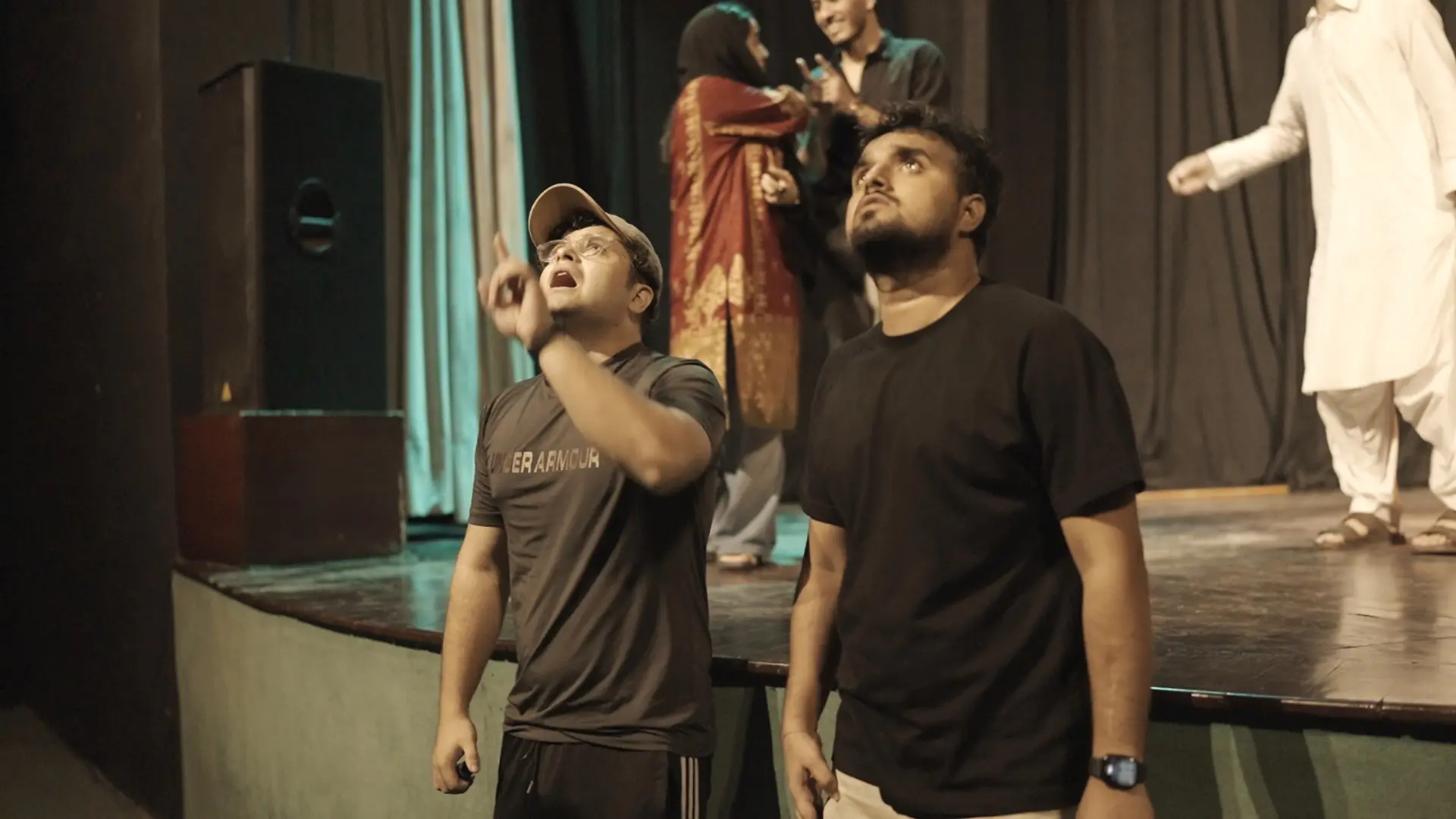 Two crew members coordinate backstage while performers continue a scene on stage behind them
