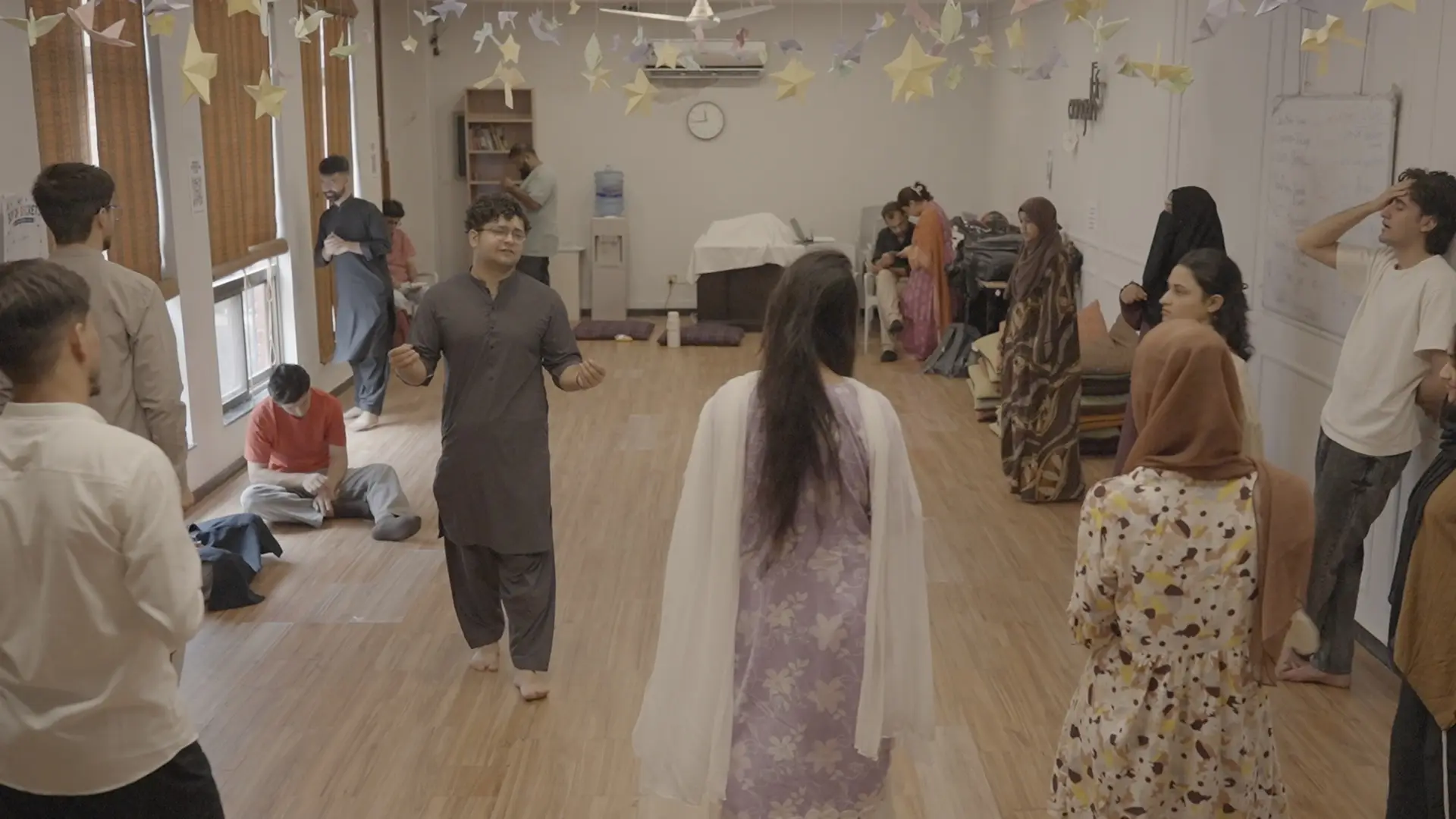 A participant moves through the centre of the hall during a theatre exercise while the cohort watches from the floor cushions