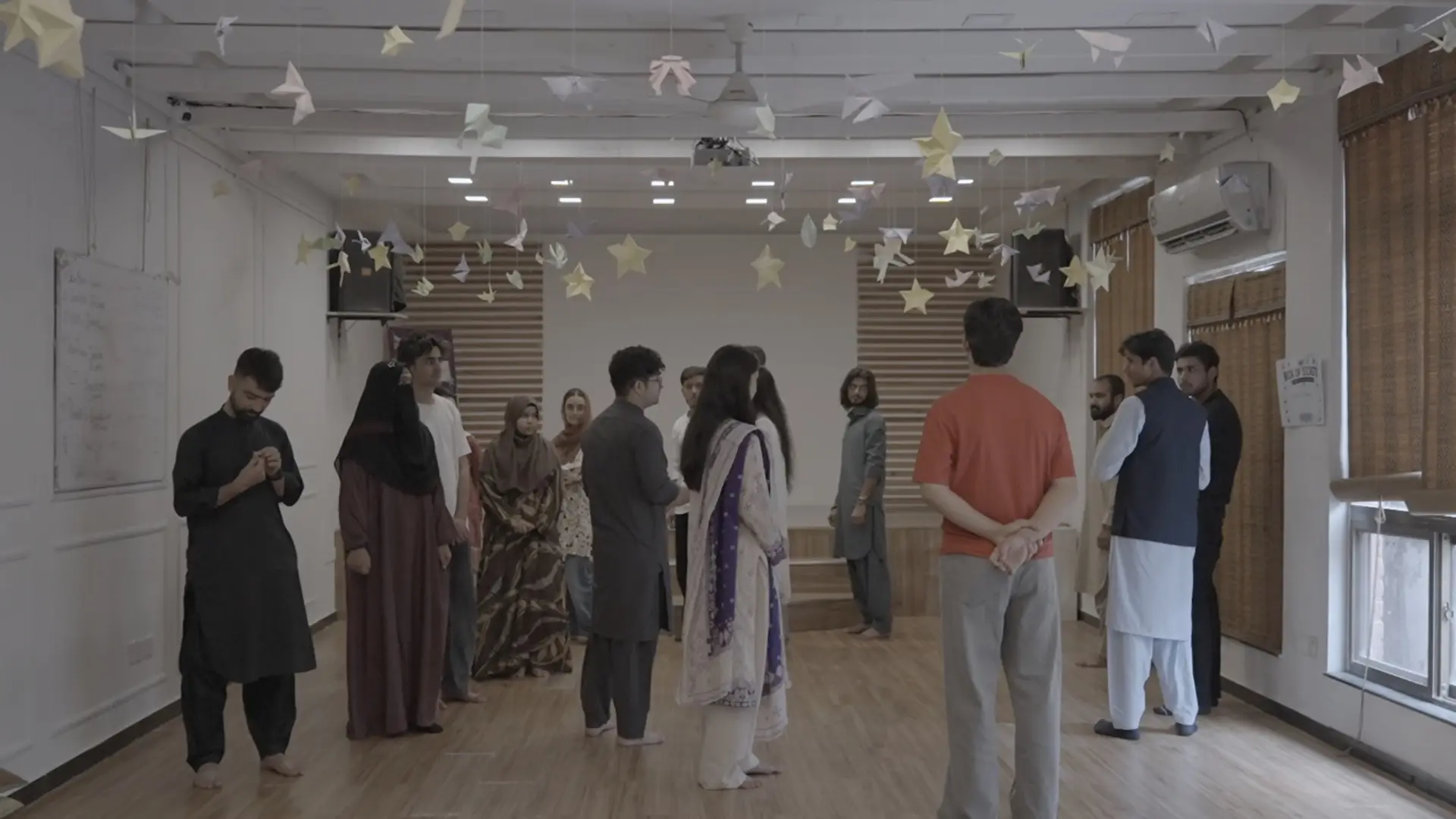 Sixteen young participants stand in a circle during a workshop at Aangun, under paper stars suspended from the ceiling